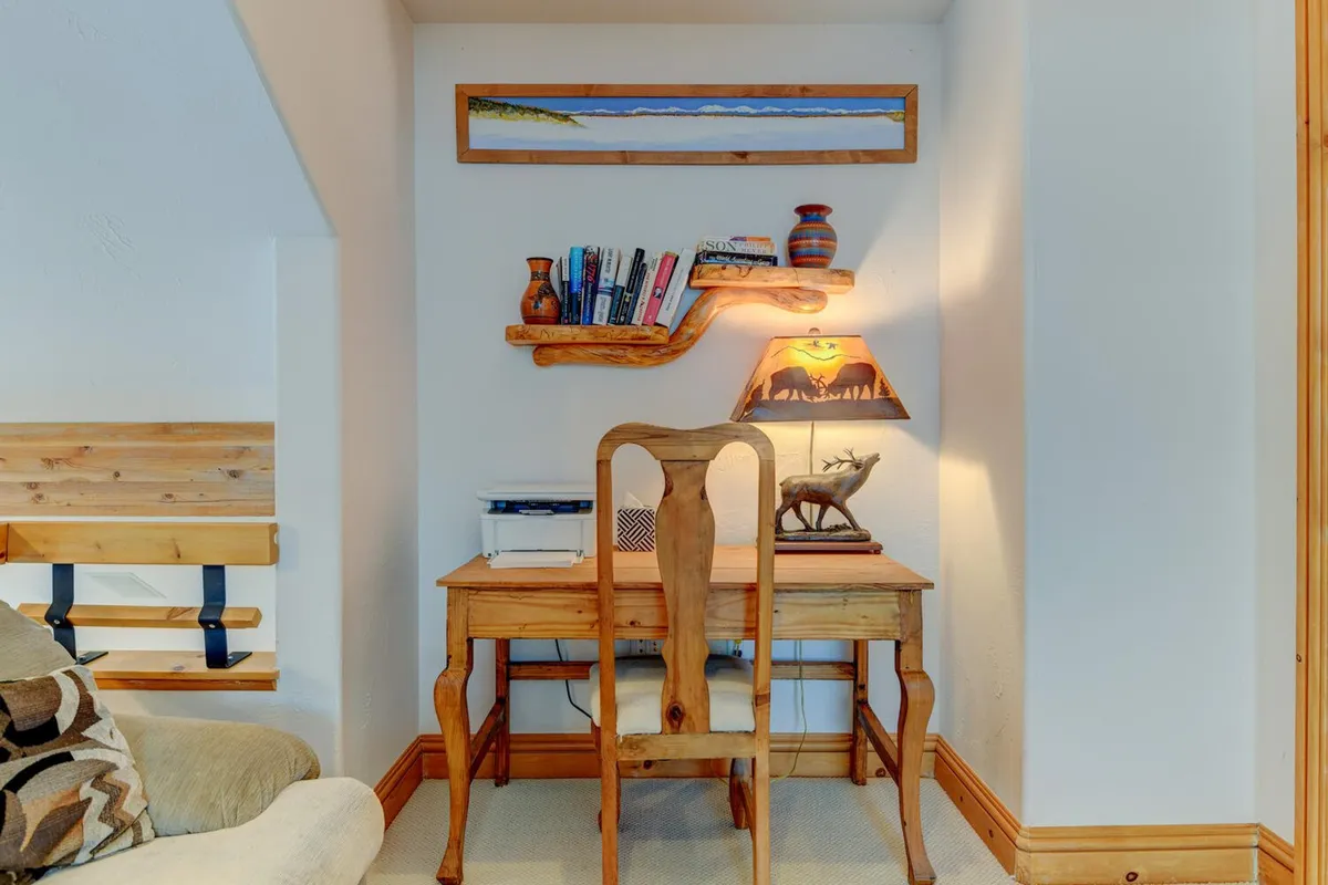A tranquil workspace with a wooden desk, bookshelf filled with books, a decorative lamp, and artwork on the wall. The setting feels peaceful and organized, inviting both productivity and creative work.