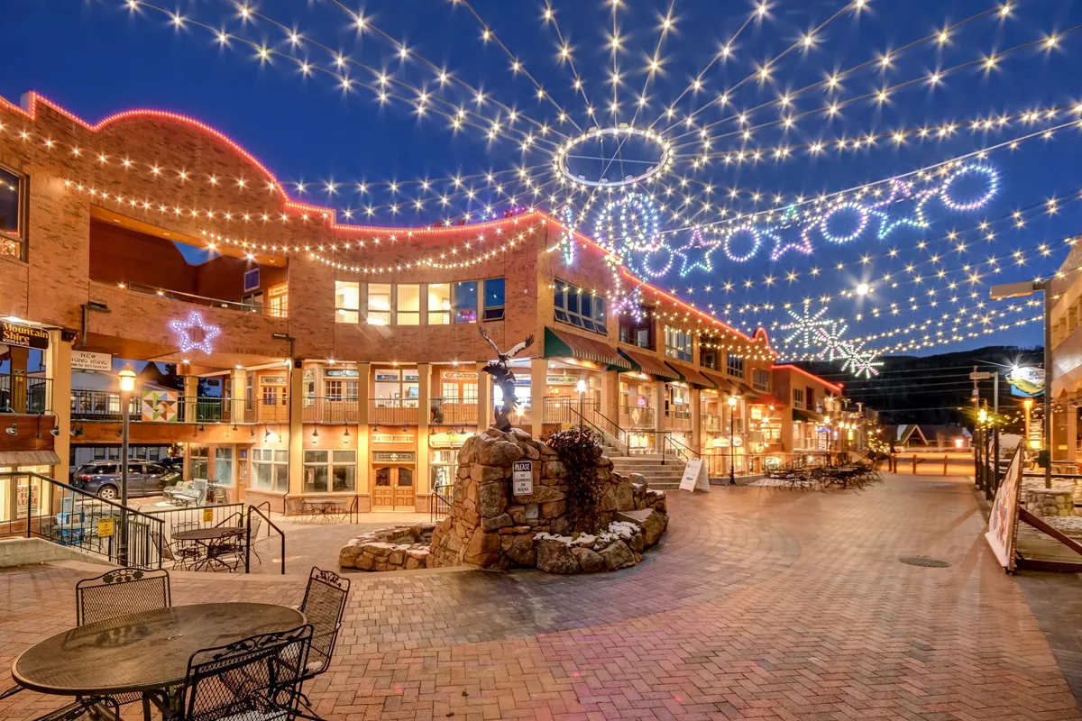 Twinkling string lights illuminate decorated storefronts and a central stone fountain, creating a festive and welcoming ambiance. People can be seen enjoying evenings out, dining, shopping, and strolling under the lights, reinforcing the sense of community and local celebration.
