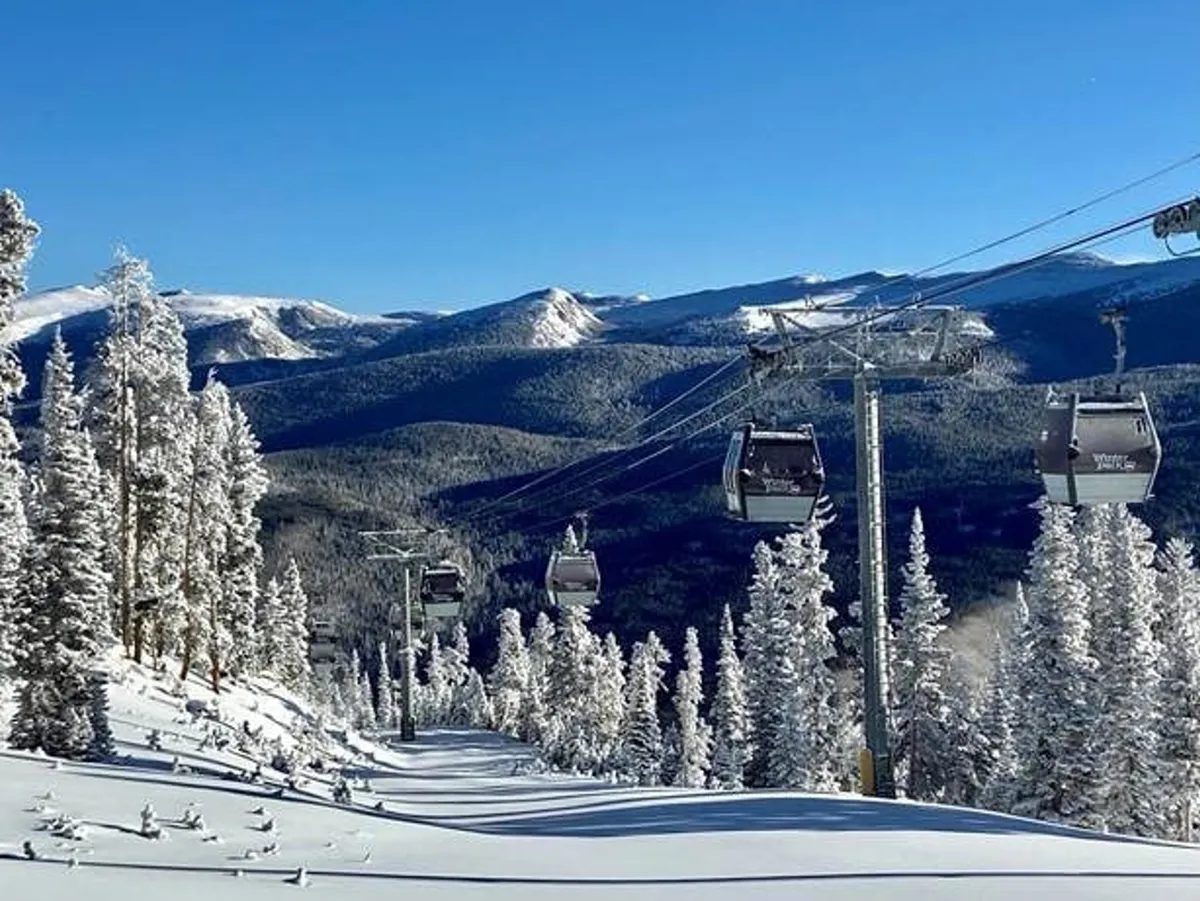 A striking exterior view of a ski lift gliding above snow-covered trees with the mountain range and a clear blue sky in the background. The landscape is blanketed in pristine snow, capturing the excitement and beauty of winter sports and outdoor adventure.