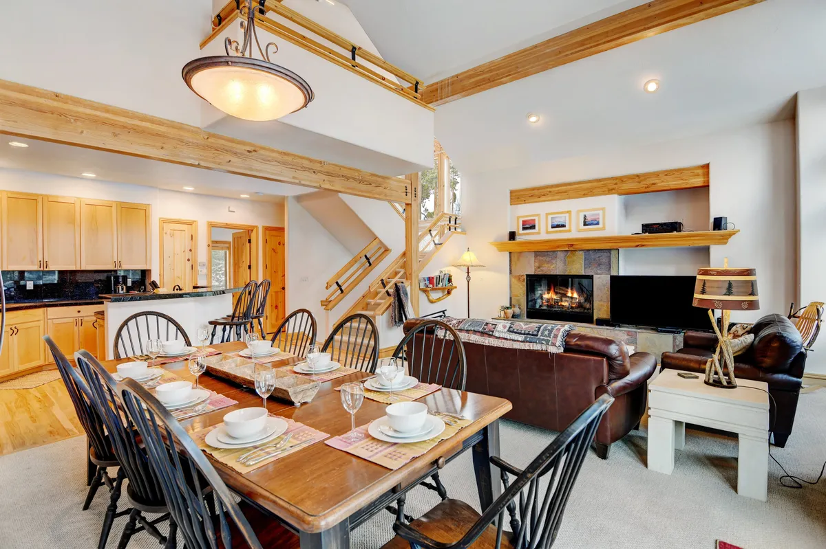 An inviting dining area set for a meal, adjacent to a fireplace with a staircase leading to an upper level. The dining table is large enough for family gatherings or entertaining friends, and the overall atmosphere is warm and festive, suitable for group meals or board games.