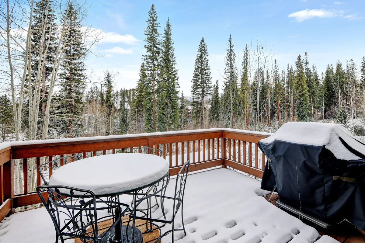 A snow-dusted private deck surrounded by tall pines, featuring a metal bistro table and a grill. The atmosphere is peaceful and distinctly mountain-like, providing a seamless extension of the indoor gathering spaces to the outdoors.
