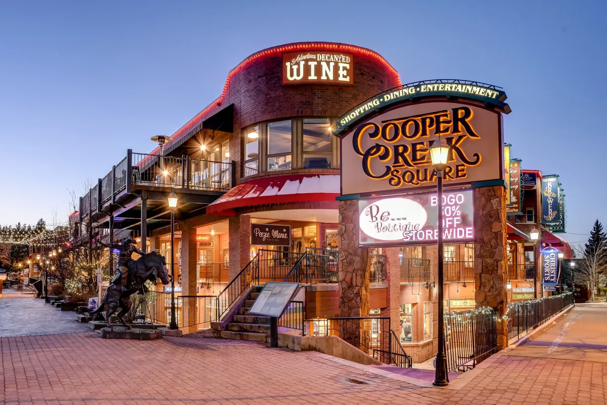 The photo shows the bustling Cooper Creek Square, with its recognizable sign, an inviting wine shop, outdoor dining spaces, and a striking horse sculpture. The area buzzes with activity, suggesting opportunities for boutique shopping, socializing, and enjoying local cuisine.