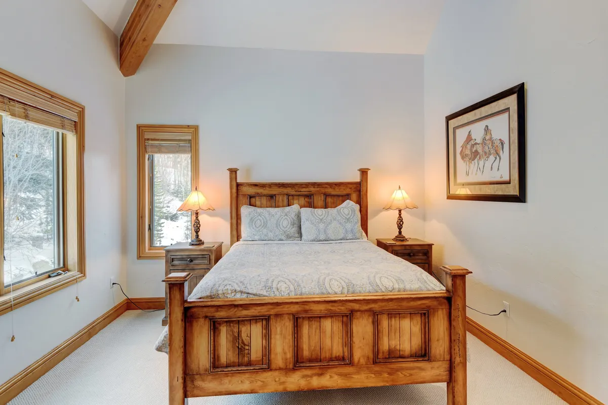 A tranquil bedroom featuring a wooden bed frame, decorative lamps on either side, tasteful artwork on the wall, and a large window letting in morning light. The overall mood is cozy and inviting, emphasizing comfort and privacy.