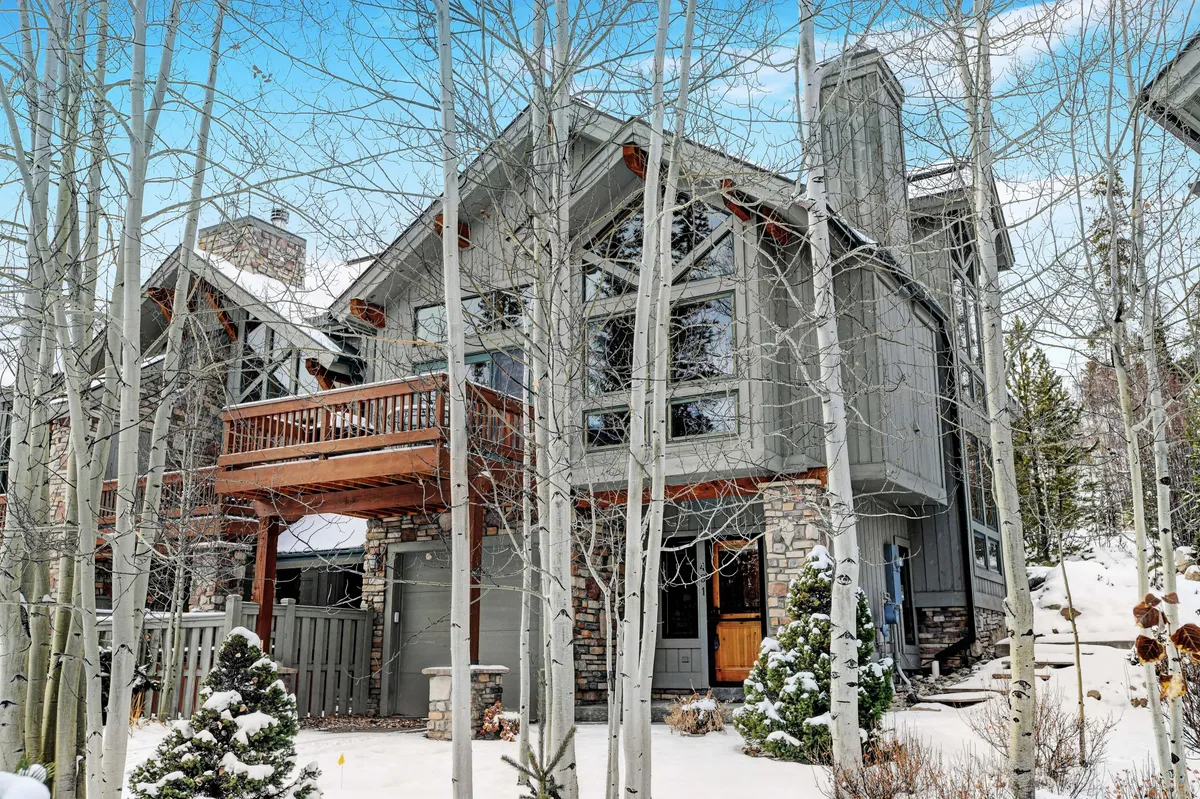 The image showcases the exterior of a luxurious single-family house nestled in a snow-covered landscape. The home features expansive glass windows, a wooden balcony perfect for taking in panoramic mountain views, and an elegant stone facade. The cozy, inviting atmosphere is highlighted by hints of activity, such as space for gathering after winter sports or enjoying hot drinks outdoors while surrounded by nature.
