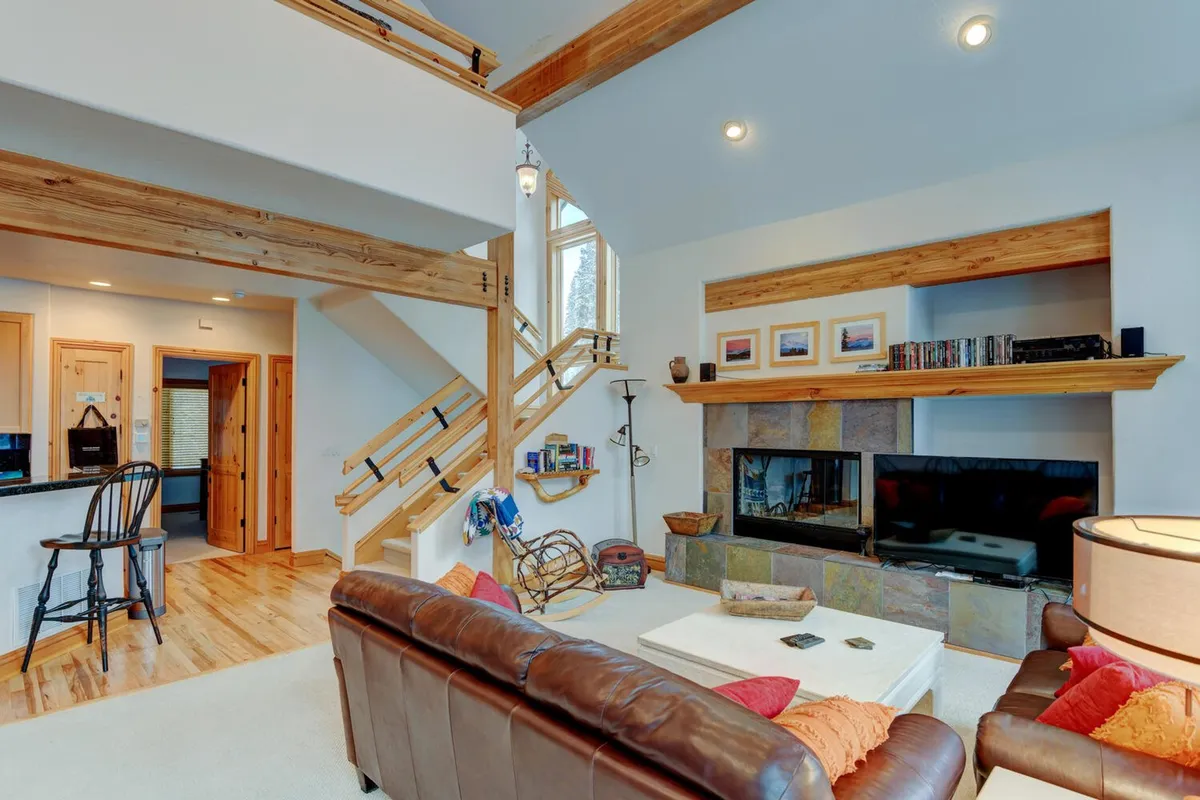 A spacious open living room centered around a large leather sofa and a glowing fireplace. The room features a prominent wooden staircase, decorative shelving, and large windows that hint at scenic mountain views. The seating area is arranged for comfortable gatherings, with a TV visible for entertainment.